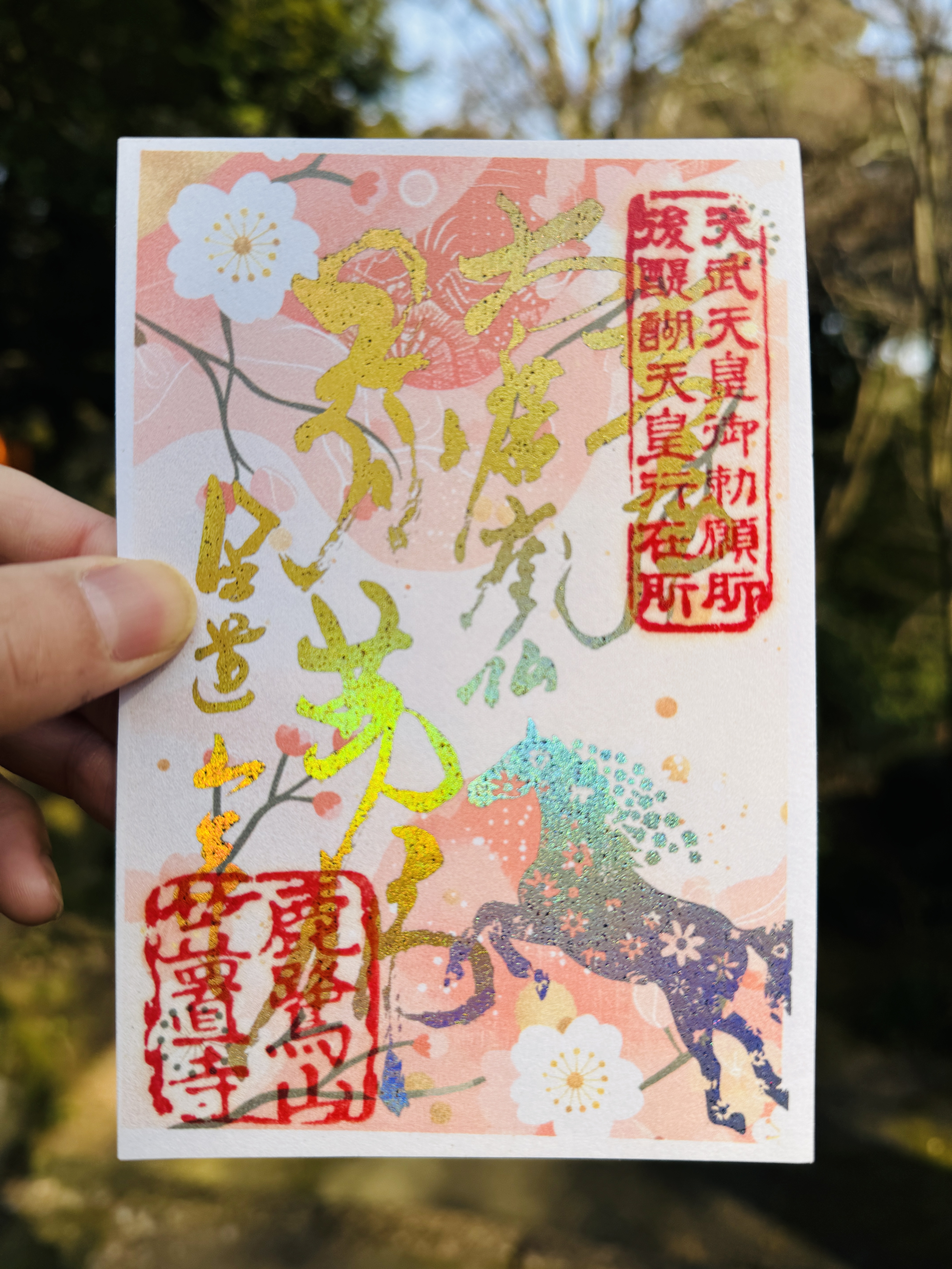 笠置寺の1月限定御朱印。淡いピンクの梅の花が描かれた背景に、金色の箔押し文字と、虹色に光る干支の馬があしらわれたデザイン。