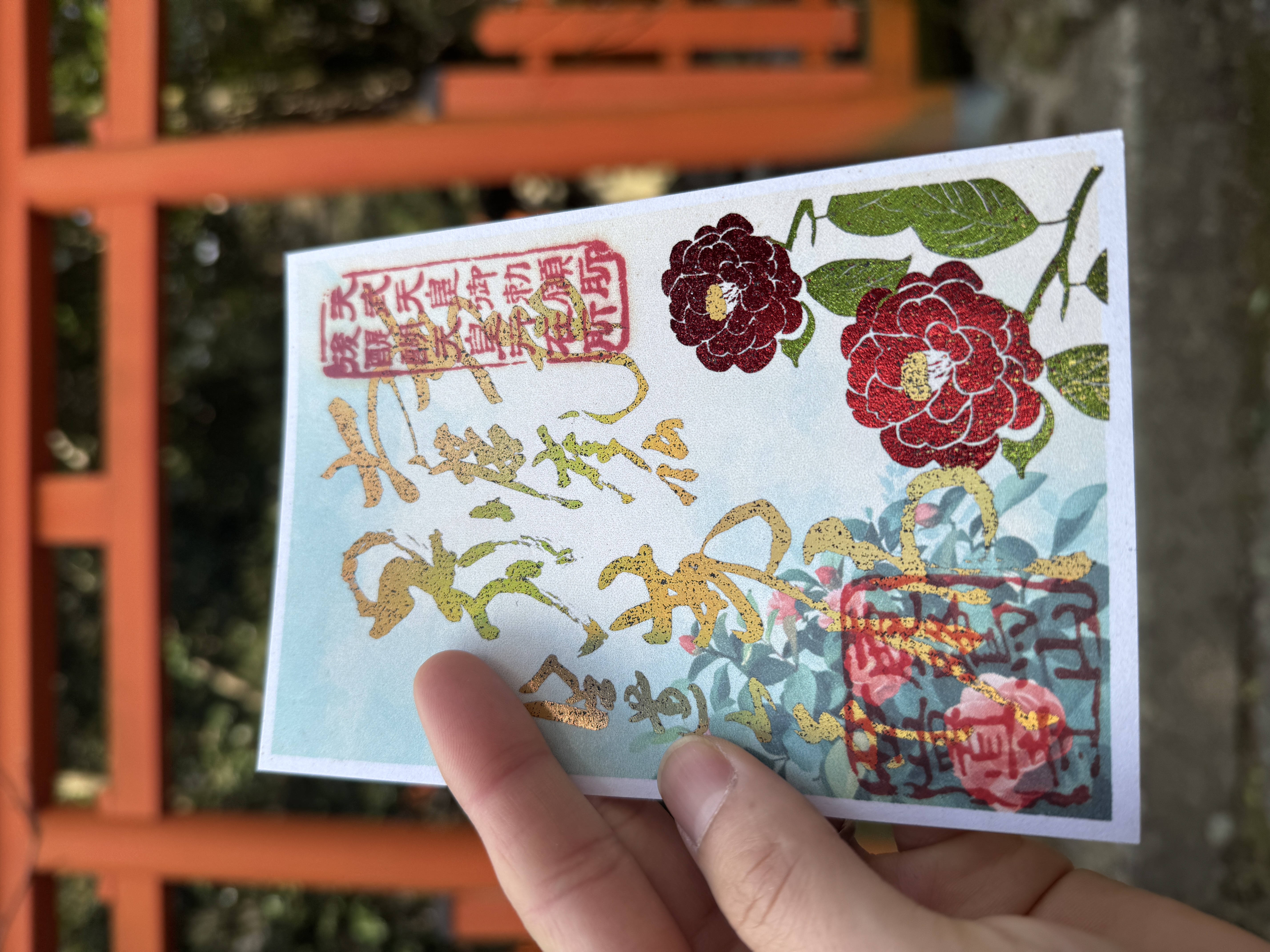 笠置寺の2月限定御朱印。鮮やかな赤い椿の花と緑の葉が描かれ、中央には金色の箔押し文字、背景には朱色の鳥居が写っている。